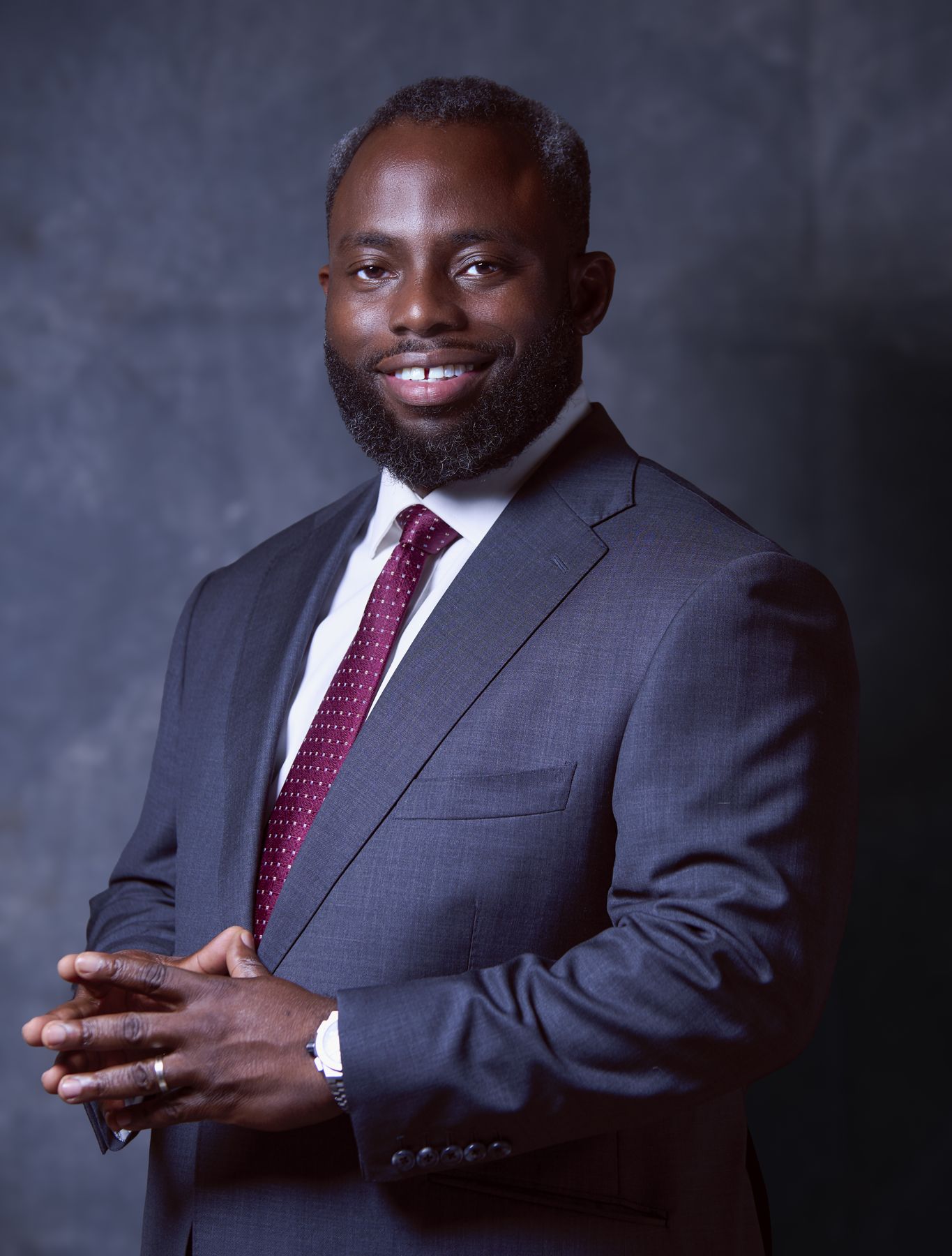 Dr Olamide Okulaja in a navy suit and burgundy tie, hands clasped